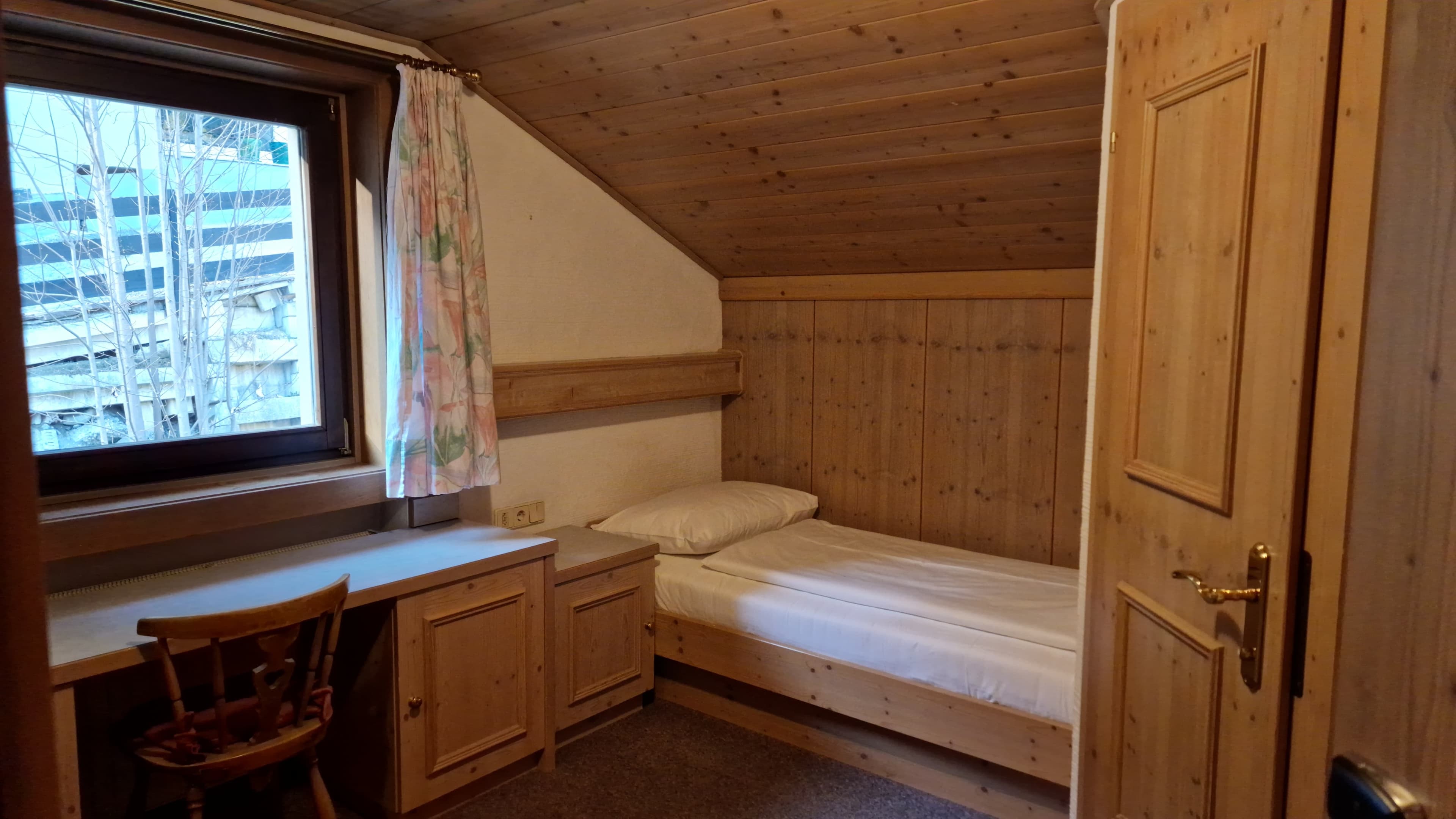 Pension Auwirt · Einzelzimmer - A cozy bedroom with a single bed made with white linens. A wooden desk and chair are placed near a window with floral curtains. The room features wooden paneling on the walls and ceiling, creating a rustic ambiance.